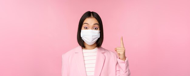 Cierra el retrato de una mujer de negocios asiática con mascarilla médica y traje señalando con el dedo hacia arriba mostrando la pancarta superior del anuncio de pie sobre fondo rosa