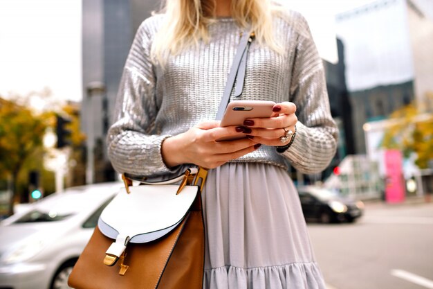 Cierre los detalles de moda de la ciudad de una mujer elegante y elegante con suéter plateado, falda de seda, bolso de cuero de lujo y gafas de sol, posando en la calle de Nueva York cerca de los centros de negocios, toque su teléfono.