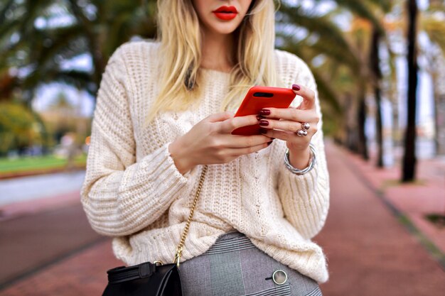 Cierre los detalles de la mujer posando en la calle y toque en su teléfono inteligente, labios rojos y suéter blanco de moda acogedor, moda.