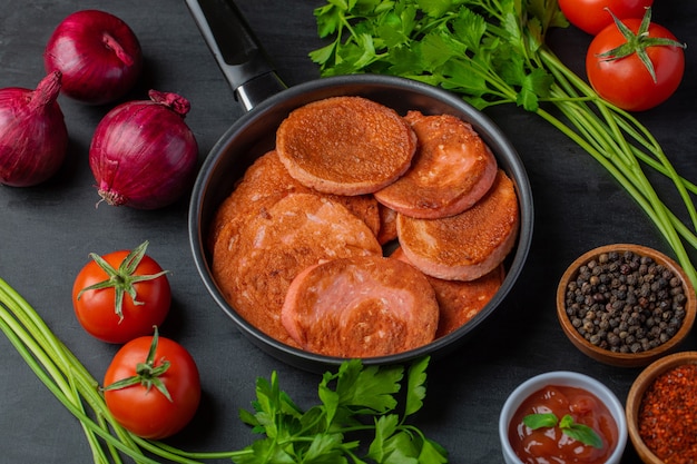 Cierre la foto de rodajas de salami frito en la sartén y varios tipos de verduras alrededor.