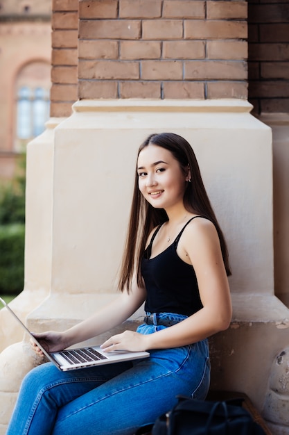 Ciérrese para arriba de un estudiante universitario de sexo femenino que se sienta en un césped usando su computadora portátil en un día soleado.