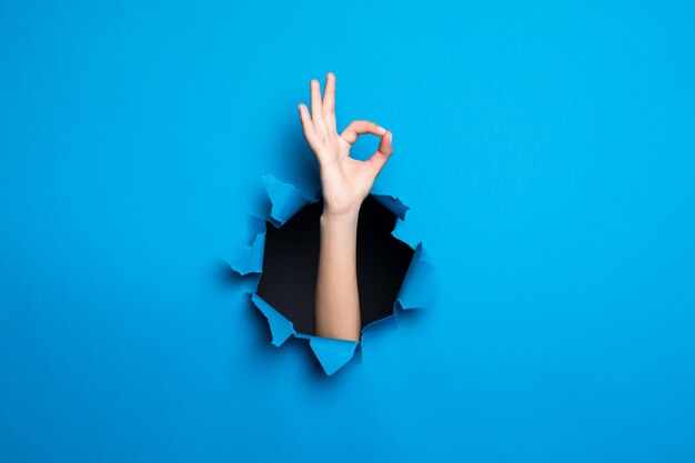 Ciérrese para arriba de la mano de la mujer con gesto aceptable a través del agujero azul en la pared de papel.