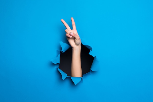 Ciérrese para arriba de la mano de la mujer con gesto de paz a través del agujero azul en la pared de papel.