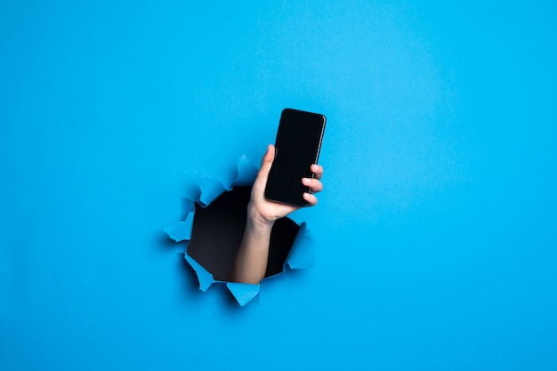 Ciérrese para arriba de la mano de la mujer que sostiene el teléfono con screan para el adv a través del agujero azul en la pared de papel.