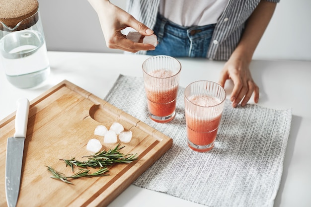 Ciérrese para arriba de las manos de la mujer que ponen pedazos del hielo en vidrios con el smoothie sano de la desintoxicación de la toronja.