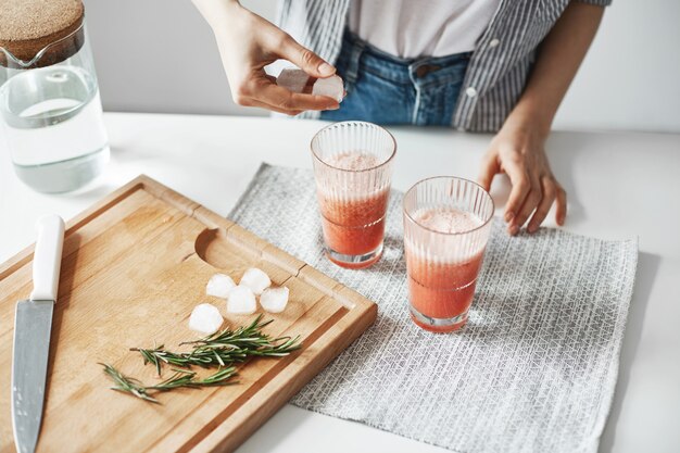 Ciérrese para arriba de las manos de la mujer que ponen pedazos del hielo en vidrios con el smoothie sano de la desintoxicación de la toronja.