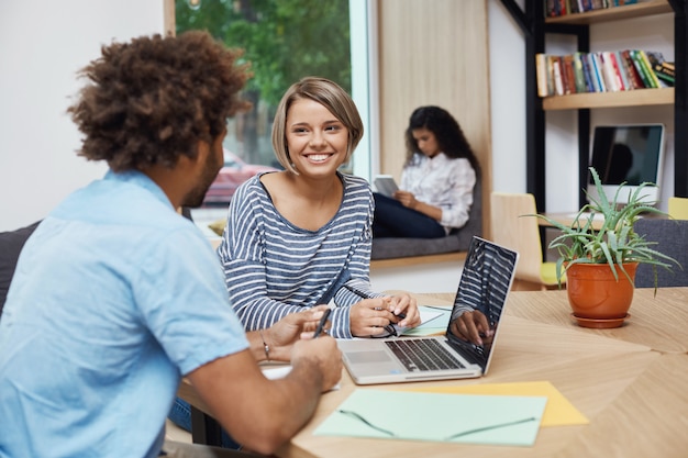 Foto gratuita ciérrese para arriba de la muchacha alegre joven del estudiante con el pelo claro en el peinado de la sacudida que se sienta en la reunión con el amigo de la universidad, haciendo proyecto del equipo, buscando la información en la computadora portátil.