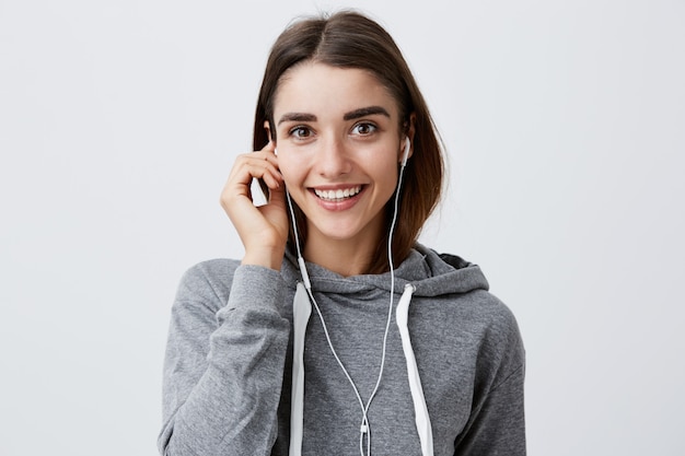 Ciérrese para arriba de la muchacha caucásica morena atractiva joven alegre en sudadera con capucha gris casual que sonríe brillantemente, sosteniendo el auricular con la mano, con la expresión feliz y relajada de la cara.