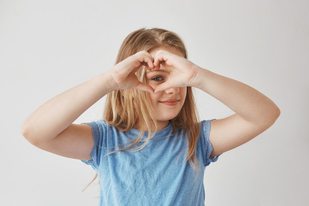 Ciérrese para arriba de la muchacha rubia hermosa que hace el corazón con las manos, mirando a través de él, posando para la foto con sonrisa y expresión feliz.