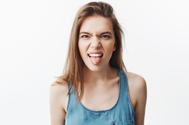 Ciérrese para arriba de la mujer joven caucásica apuesta tonta con el pelo oscuro en la camisa azul que muestra la lengua, haciendo la expresión de la cara mala, mostrando falta de respeto al pequeño niño durante el juego.