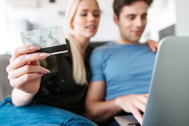 Ciérrese para arriba de la mujer joven que sostiene la tarjeta de crédito mientras que usa la computadora portátil con su marido