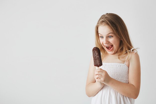 Ciérrese para arriba de la pequeña muchacha rubia linda con los ojos azules que miran el helado en sus manos con la boca abierta, emocionado por la comida malsana.