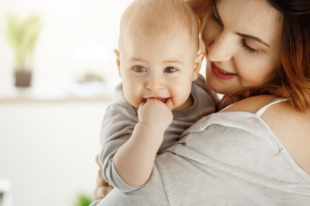 Ciérrese para arriba de pequeño bebé lindo en las manos de la madre. Mamá mirando al niño con amor y cuidado mientras el niño royendo la mano y sonriendo a la cámara. Concepto de familia