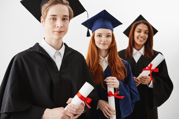 Ciérrese para arriba de tres graduados universitarios internacionales felices de la raza mixta que sonríen alegres que sostienen los diplomas. Futuros abogados o cirujanos.