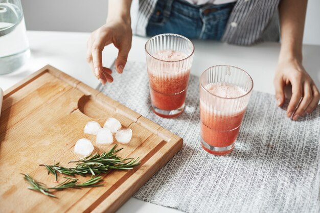 Ciérrese para arriba de los vidrios de las manos de la mujer con los pedazos del romero y del hielo del smoothie de la dieta de la desintoxicación del pomelo en el escritorio de madera.