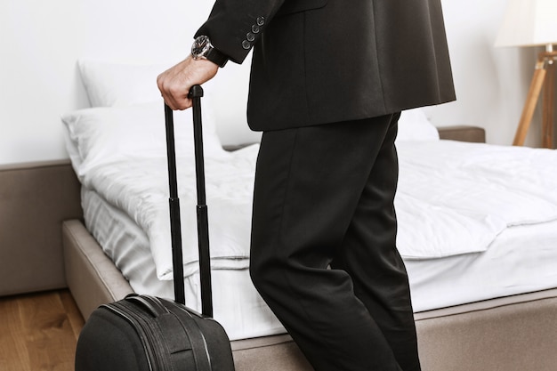 Ciérrese encima del detalle del hombre de negocios elegante en el traje negro que sostiene la maleta en las manos que van a salir de la habitación de hotel y volar a casa en avión de viaje de negocios.