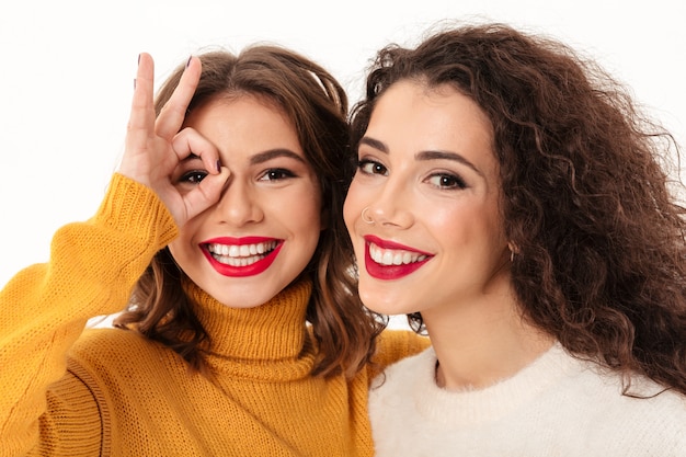 Ciérrese encima de dos muchachas felices en suéteres que se divierten sobre la pared blanca