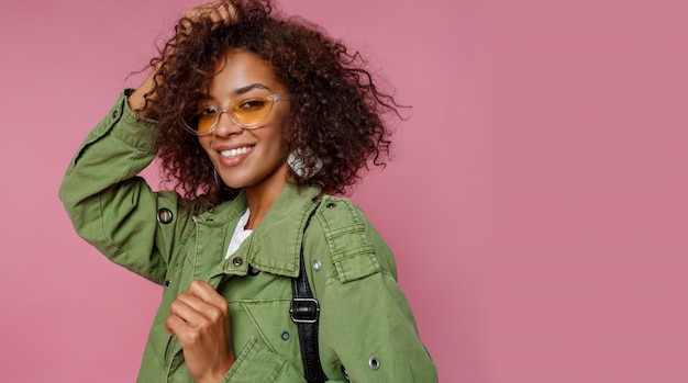 Ciérrese encima de la foto de la muchacha africana rizada asombrosa en fondo rosado. Vistiendo chaqueta verde de moda.