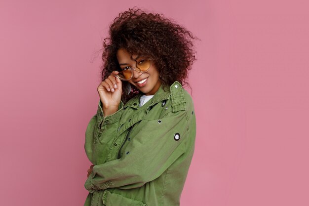 Ciérrese encima de la foto de la muchacha africana rizada asombrosa en fondo rosado. Vistiendo chaqueta verde de moda.