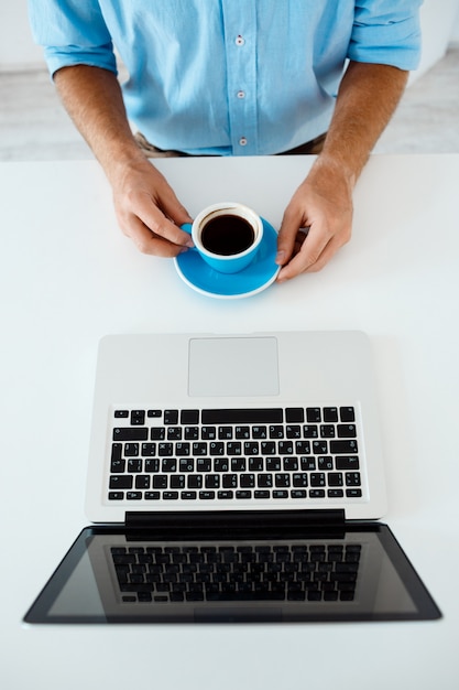 Ciérrese encima de la imagen de las manos del hombre de negocios joven que se sientan en la tabla que trabaja en la computadora portátil que sostiene la taza de café. Interior de oficina moderno blanco