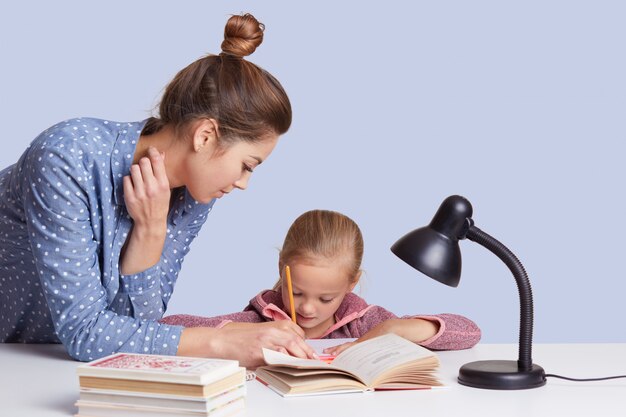 Ciérrese encima de la mamá joven ayuda a escribir a su hija para escribir la composición, utilice la lámpara de lectura, las muchachas parecen concentradas, aisladas en blanco. Los niños y el concepto de aprendizaje.