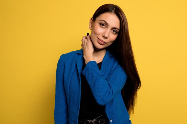 Ciérrese encima de la mujer encantadora adorable en la chaqueta azul que presenta con la sonrisa sobre la pared amarilla. Modelo femenino europeo posando