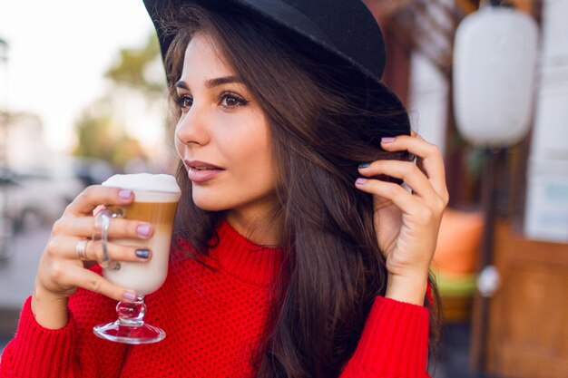 Ciérrese encima de mujer morena joven hermosa en sombrero negro elegante y suéter rojo brillante que se sienta en café del espacio abierto y beba el café con leche o capuchino en mañana soleada.