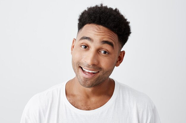 Ciérrese encima del retrato aislado del joven estudiante masculino alegre hermoso de piel oscura con el peinado afro en la camiseta blanca casual que sonríe con los dientes, mirando en cámara con la expresión feliz y emocionada.