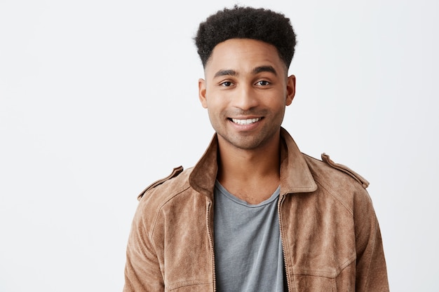 Foto gratuita ciérrese encima del retrato aislado del tipo atractivo joven de piel oscura con el peinado afro en camiseta gris debajo de la chaqueta marrón que sonríe con los dientes que miran en cámara con la expresión feliz y pacífica de la cara.