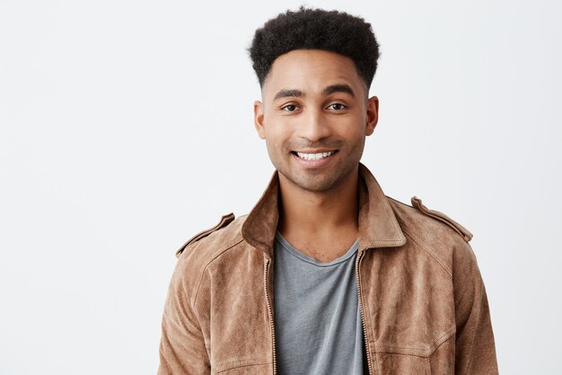 Ciérrese encima del retrato aislado del tipo atractivo joven de piel oscura con el peinado afro en camiseta gris debajo de la chaqueta marrón que sonríe con los dientes que miran en cámara con la expresión feliz y pacífica de la cara.