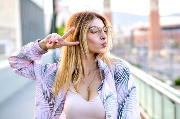 Ciérrese encima del retrato al aire libre de la mujer rubia feliz, posando en la calle, enviando besos y mostrando gesto de paz, tiempo de primavera, ropa de moda, colores suaves.