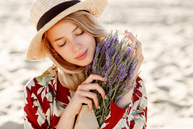 Ciérrese encima del retrato de la belleza de la muchacha rubia romántica encantadora que disfruta del olor perfecto de la lavanda. Concepto de cosmética y cuidado de la piel. Colores cálidos del atardecer. Ojos cerrados.