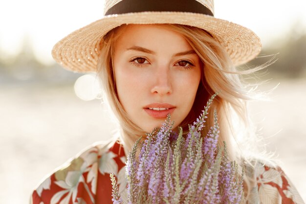 Ciérrese encima del retrato de la belleza de la muchacha rubia romántica encantadora que disfruta del olor perfecto de la lavanda. Concepto de cosmética y cuidado de la piel. Colores cálidos del atardecer.