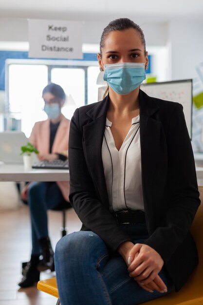 Ciérrese encima del retrato del empleado de la empresa en el espacio de trabajo con la mascarilla como precaución de seguridad durante la pandemia global con el coronavirus mirando a la cámara.