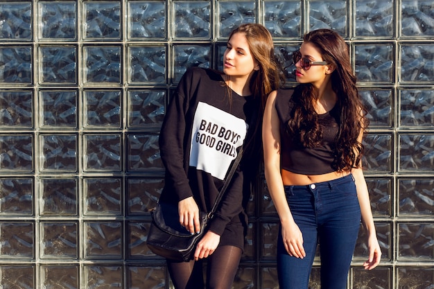 Ciérrese encima del retrato del estilo de vida de dos mujeres bastante jóvenes que presentan contra la pared urbana de cristal el día de sonny. Vistiendo blusa negra, suéter y chaqueta de cuero.