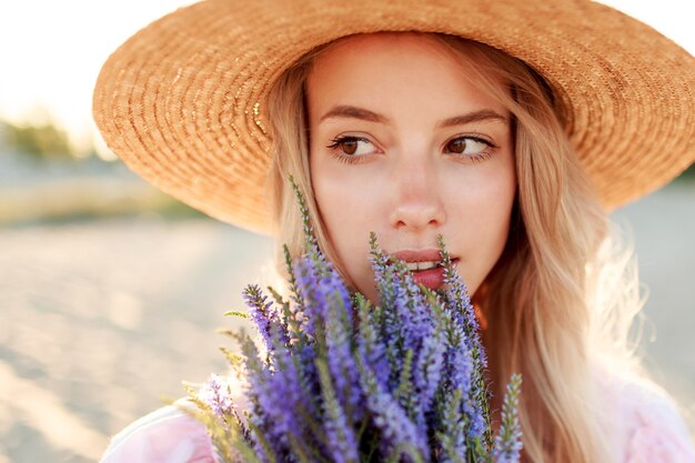 Ciérrese encima del retrato del estilo de vida de la mujer rubia romántica con las flores en la mano que camina en la playa soleada. Colores cálidos del atardecer.