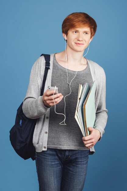 Ciérrese encima del retrato del estudiante apuesto joven alegre confidente con la mochila que sostiene los libros en manos, charlando en smartphone con el amigo, escuchando música en auriculares.