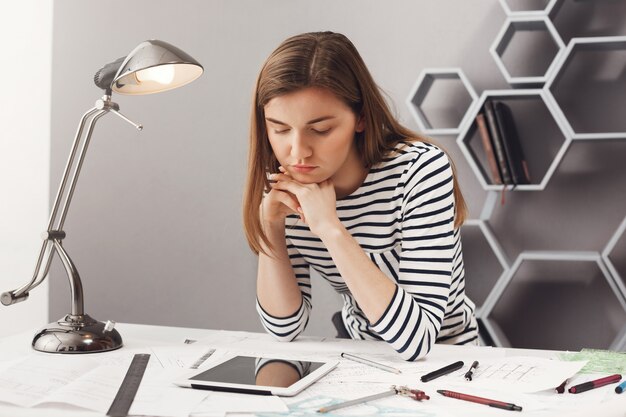 Ciérrese encima del retrato de la estudiante joven hermosa hermosa del arquitecto femenino con el pelo castaño en mirada rayada, sosteniendo la cabeza con las manos, mirando en tableta digital con expresión de la cara cansada, buscando exampl