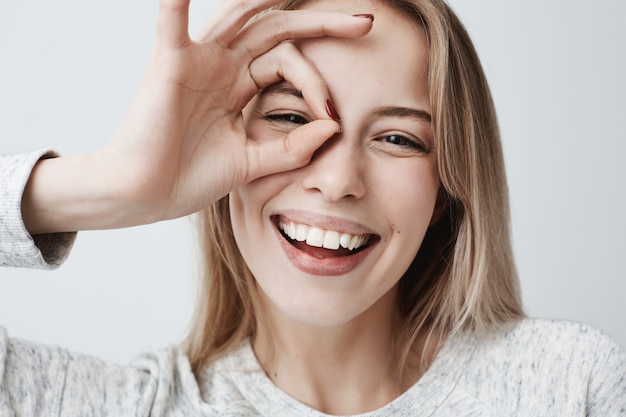 Ciérrese encima del retrato de la hembra rubia caucásica alegre hermosa que sonríe, demostrando los dientes blancos, mirando con los dedos en gesto aceptable. Expresiones faciales, emociones y lenguaje corporal.