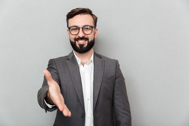 Ciérrese encima del retrato del hombre amable amistoso en lentes que mira en cámara con sonrisa sincera, ofreciendo el apretón de manos aislado sobre gris