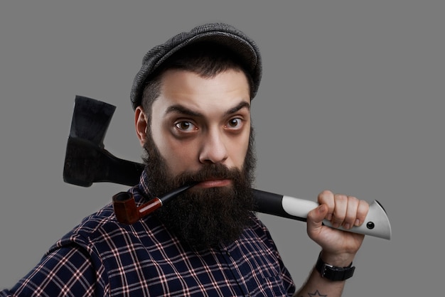 Ciérrese encima del retrato del hombre de la barba sorprendido que fuma en pipa y sostenga el hacha en la mano tatuada. Retrato masculino fuerte y serio aislado sobre fondo gris.