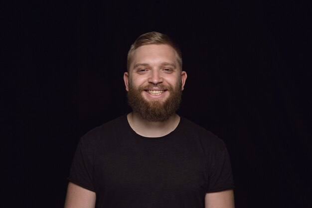 Ciérrese encima del retrato del hombre joven aislado en la pared negra. Photoshot de emociones reales del modelo masculino. Sonriendo, sintiéndose feliz. Expresión facial, concepto de emociones humanas puras y claras.