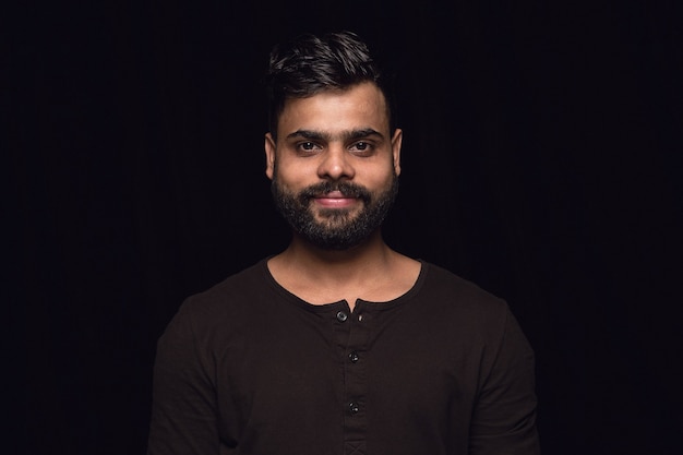 Ciérrese encima del retrato del hombre joven aislado en la pared negra. Photoshot de emociones reales del modelo masculino. Sonriendo, sintiéndose feliz. Expresión facial, concepto de emociones humanas puras y claras.