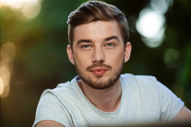 Ciérrese encima del retrato del hombre joven confiado hermoso en la camiseta blanca en la naturaleza al aire libre borrosa