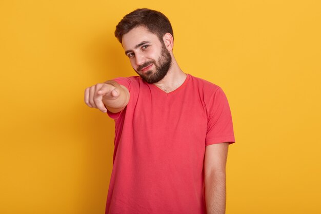 Ciérrese encima del retrato del hombre joven feliz que señala en la cámara y que sonríe mientras que, vistiendo la camiseta casual roja