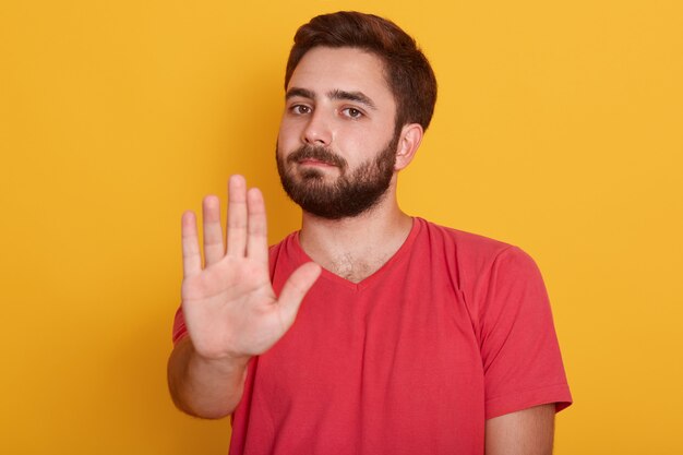 Ciérrese encima del retrato del hombre joven que requiere parar con su mano, chico guapo con camiseta roja, mostrando gesto de parada