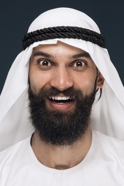 Ciérrese encima del retrato del hombre de negocios de Arabia Saudita sobre fondo azul oscuro del estudio. Modelo masculino joven de pie y sonriendo, parece feliz. Concepto de negocio, finanzas, expresión facial, emociones humanas.