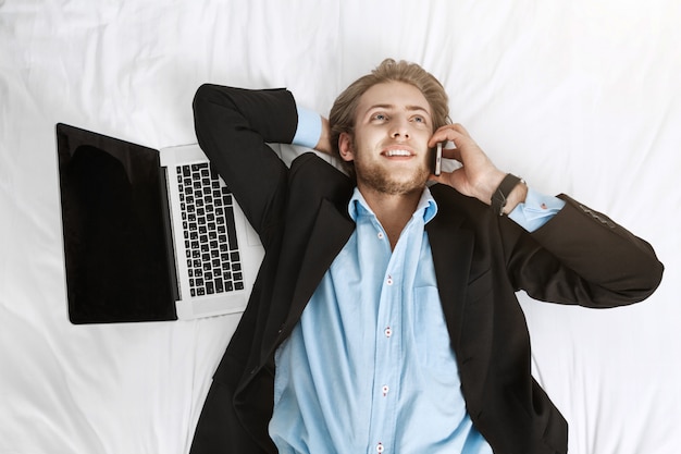 Foto gratuita ciérrese encima del retrato del hombre de negocios hermoso alegre que miente en cama en traje con la computadora portátil y el teléfono celular. hablando con el cliente, contento con su trabajo.