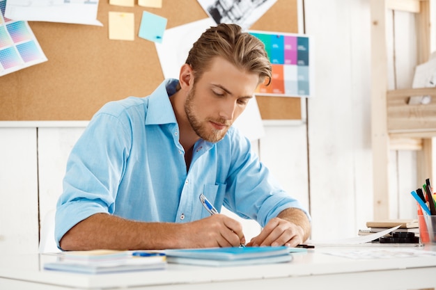 Ciérrese encima del retrato del hombre de negocios pensativo confiado hermoso joven de trabajo que se sienta en la escritura de la tabla en cuaderno. Interior de oficina moderno blanco