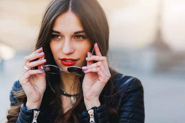 Ciérrese encima del retrato de la moda de la mujer joven bastante atractiva con las gafas de sol, posando al aire libre. Labios rojos, peinado ondulado.